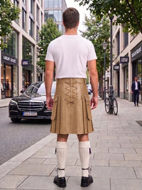 A man viewed from behind wearing a beige pleated kilt with lace-up back detailing, a white t-shirt, white knee-high socks, and black shoes, standing on a city sidewalk.