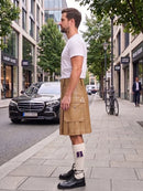 A man standing on a city sidewalk wearing a tan leather kilt, white t-shirt, white socks, and black shoes, with a street scene and parked cars in the background.