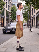 A man standing on a city sidewalk wearing a white t-shirt, a tan pleated kilt, white socks, and black leather shoes, with a street scene and parked cars in the background.