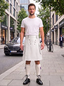 A man stands on a city sidewalk wearing a white short-sleeve t-shirt and a pleated white kilt with silver buttons, paired with white knee-high socks and black leather shoes. The background shows a street scene with buildings, a black Mercedes-Benz car, and a bicycle.