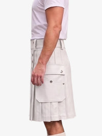 A side view of a person wearing a light beige pleated kilt with flap pockets and silver snap buttons, paired with a white t-shirt, against a plain white background.