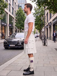 A man wearing a white short-sleeve t-shirt and a cream-colored pleated kilt stands on a city sidewalk next to a street with a black Mercedes-Benz car and trees.