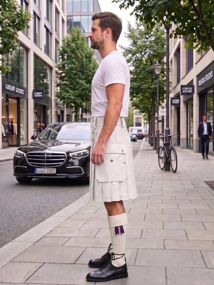 A man wearing a white short-sleeve t-shirt and a cream-colored pleated kilt stands on a city sidewalk next to a street with a black Mercedes-Benz car and trees.