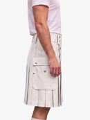 A person wearing a light beige pleated kilt with side pockets and decorative metal rivets, paired with a pale pink t-shirt, shown from the side against a plain white background.