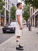 A man standing on a city sidewalk wearing a white short-sleeve t-shirt and a pleated white kilt with multiple utility pockets, paired with white socks and black leather shoes. A black Mercedes-Benz car and a bicycle are parked in the background of the street scene.