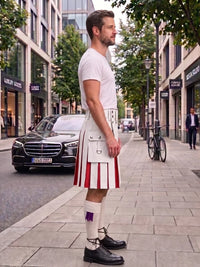 A man standing on a city sidewalk wearing a white kilt with red and white pleated detailing, a white shirt, white socks, and black shoes, with buildings, trees, and parked cars in the background.